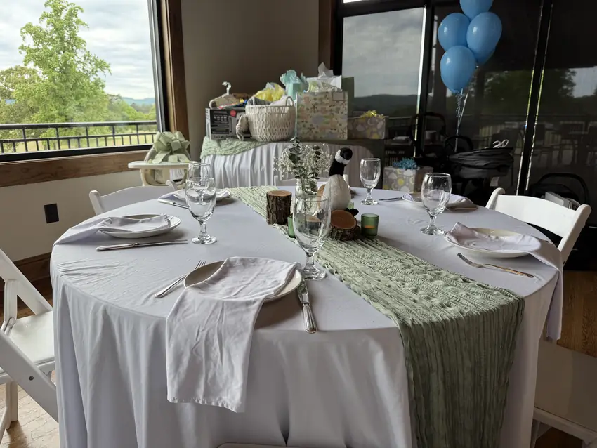 Baby shower table with sage runner, gifts, and blue balloons beside the windows