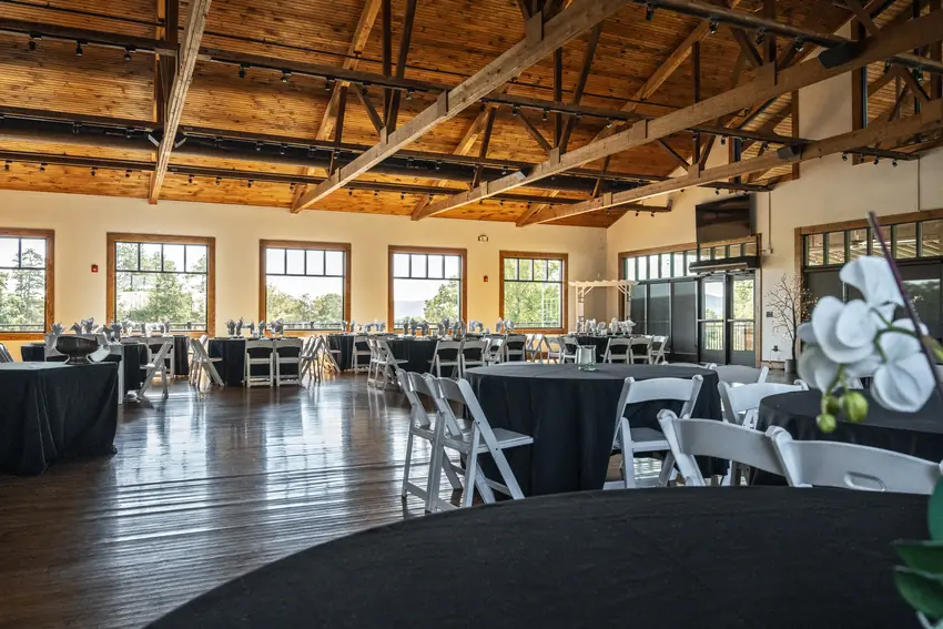 Banquet room set with round tables and white folding chairs beneath timber trusses